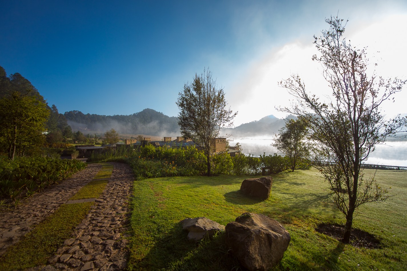 Jardín con sendero de piedra volcánica, lago y niebla matutina, Valle de Bravo
